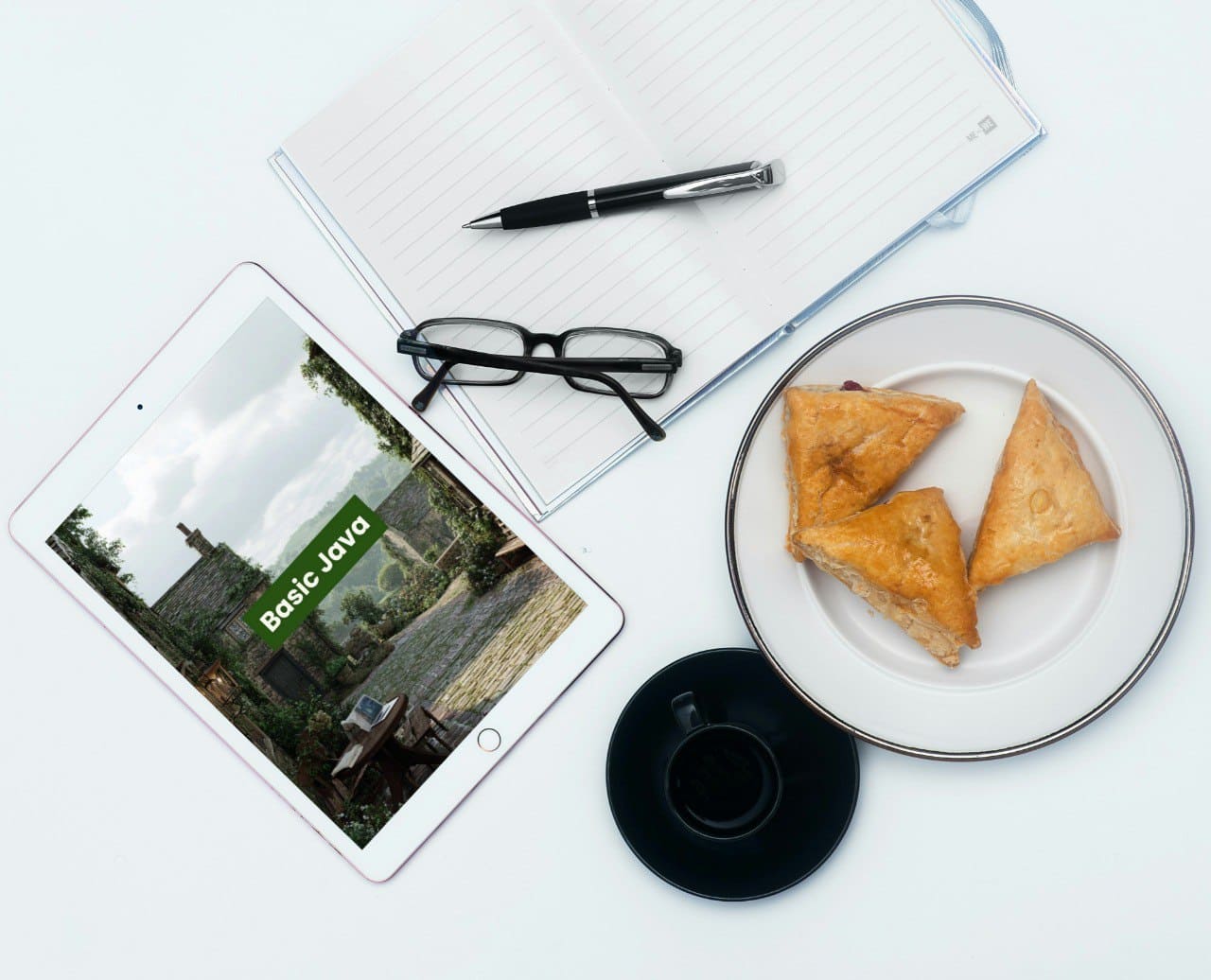 Tablet displaying a Java PDF course 'Basic Java', plate with pastries, pen, glasses, and coffee cup on a white surface 