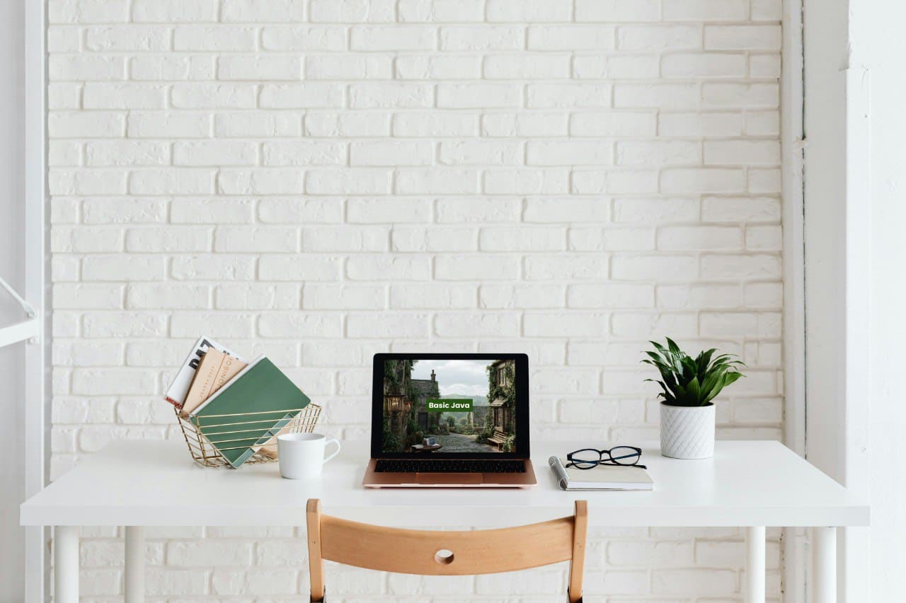Minimalist home office with a white desk, laptop displaying Java PDF course 'Basic Java', mug, plant, and chair against a white brick wall.