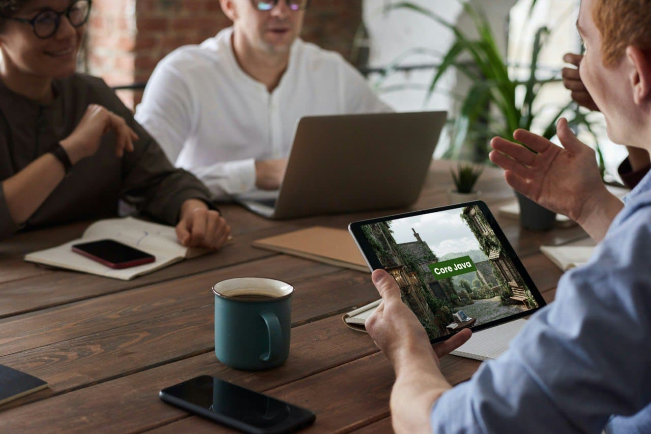 People in a meeting with a tablet and laptop displaying Java PDF course 'Core Java' on a wooden table.