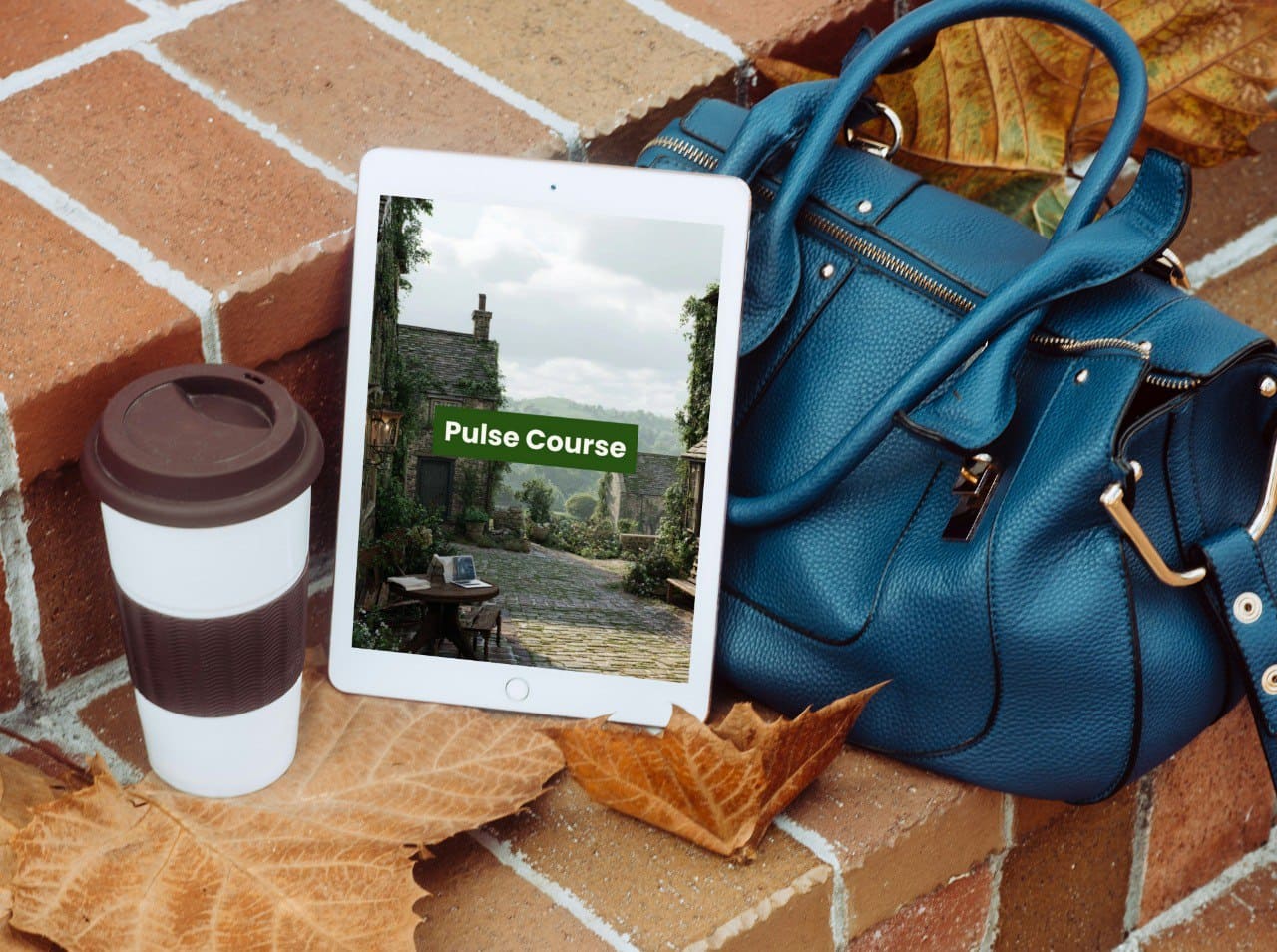 Blue handbag, white tablet displaying a course screen, and coffee cup on a brick surface with leaves.